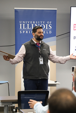 Man speaking at a presentation, University of Illinois Springfield banner behind him.
