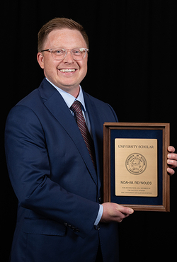 Man in suit holding a wooden plaque, smiling against a black background.