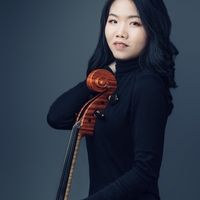 Person in black holding a cello, with a dark background.