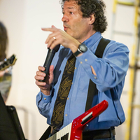 Ron Silver in blue shirt playing keytar and speaking into a microphone.