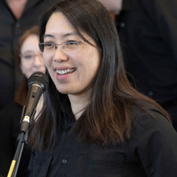 Smiling woman speaking at a microphone, wearing glasses.