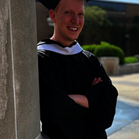 Graduate in cap and gown standing by a column.