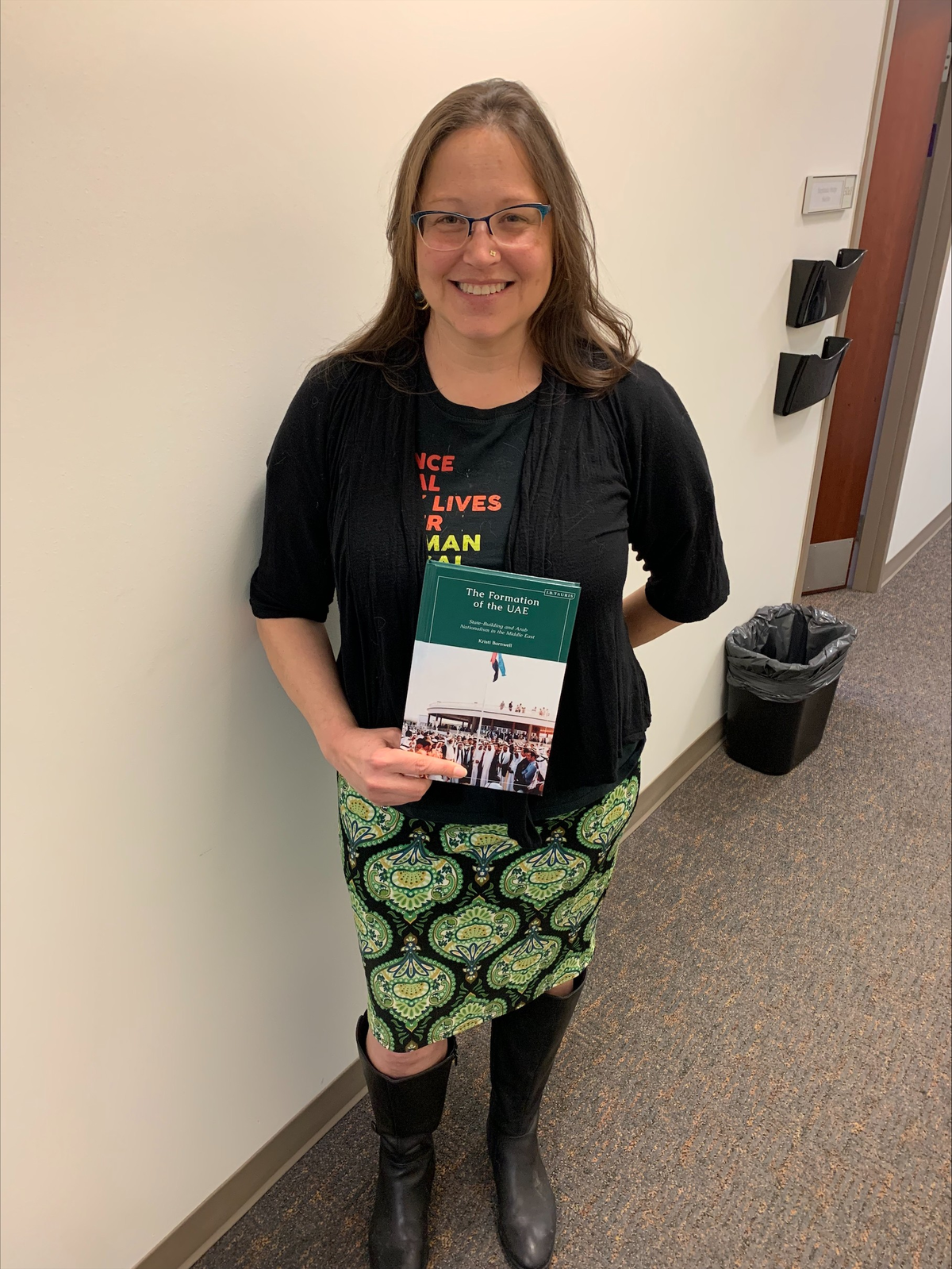 Dr. Kristi Barnwell, a white woman in a black sweater and green dress, smiling and holding a copy of her new book. 