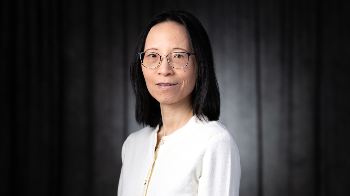 Woman with glasses, wearing a white top against a dark backdrop.