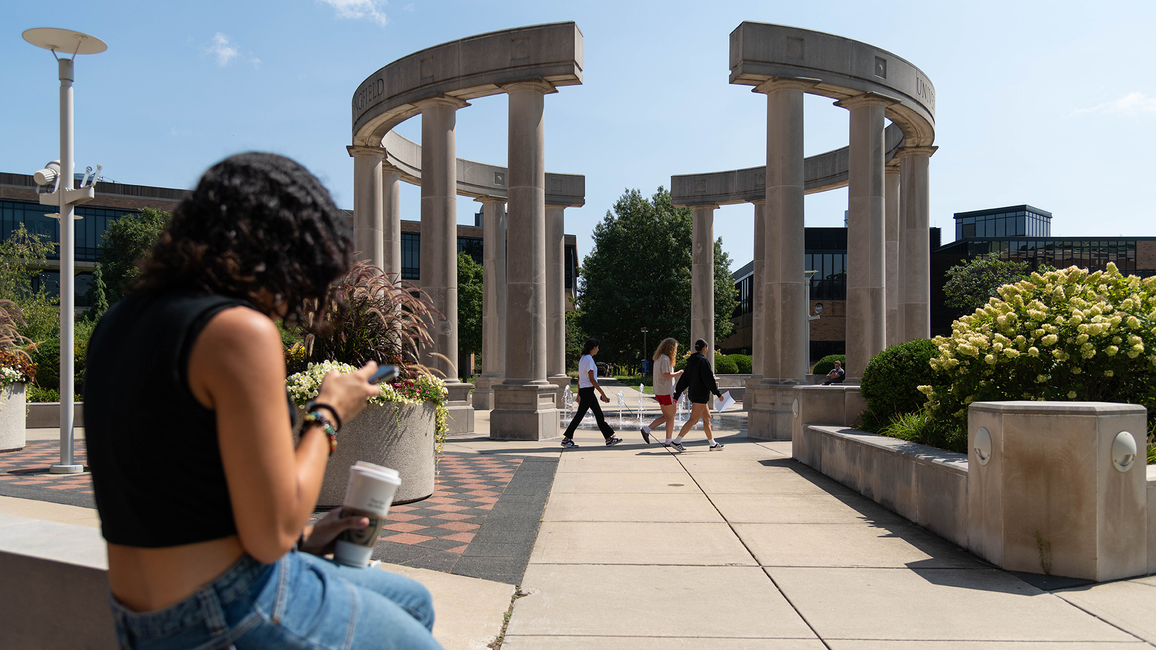 Students walk through the colonnade at the University of Illinois Springfield, while one sits nearby using a phone.