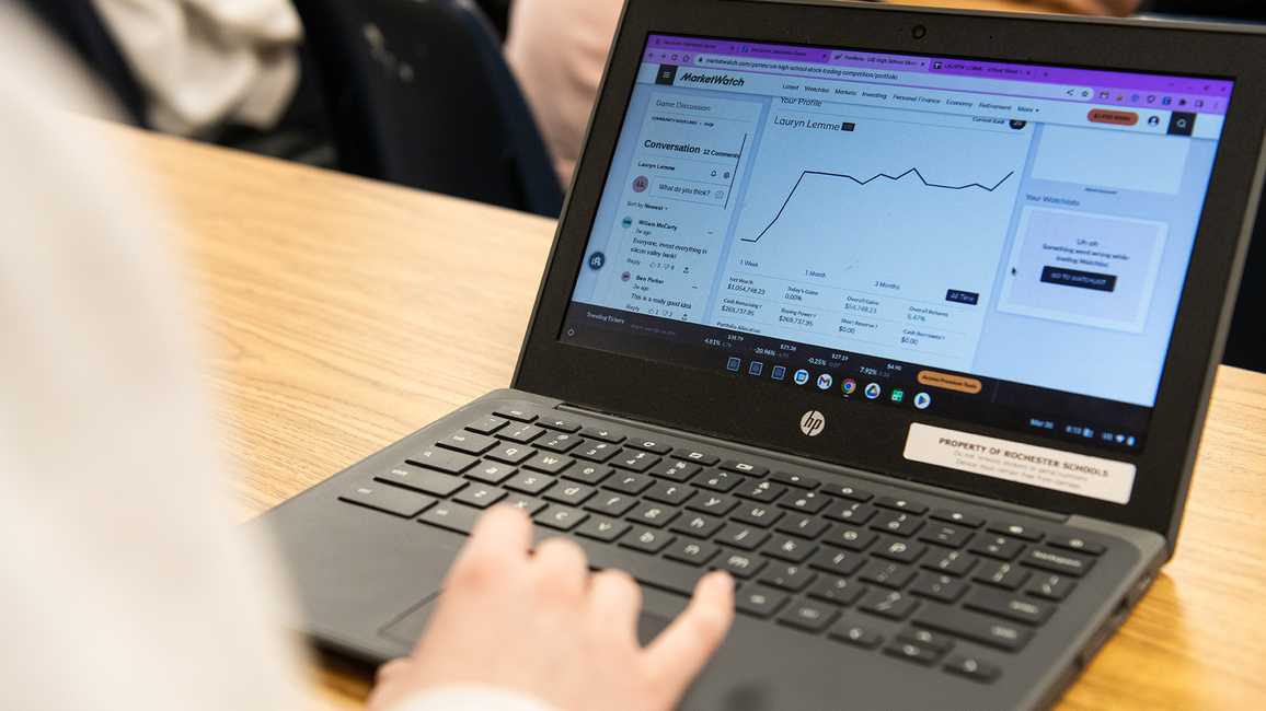 A laptop with stock trading information on the screen sitting on a desk with a person's hand above the mousepad. 