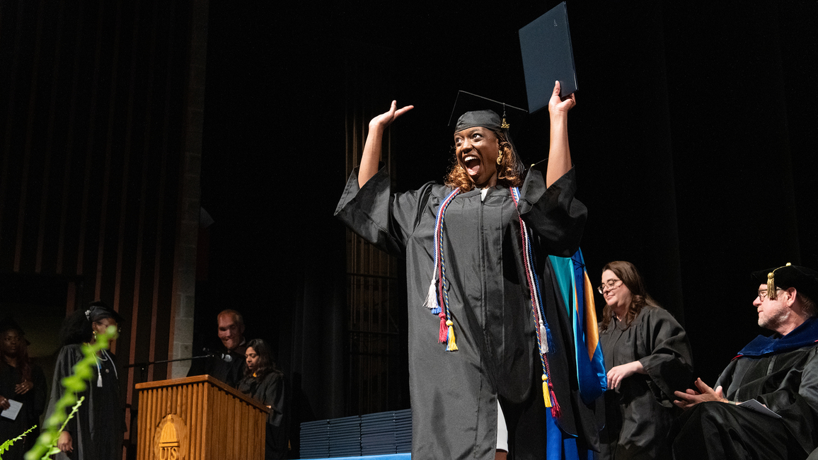 Graduating student celebrates on stage in cap and gown holding diploma at UIS Commencement as faculty applaud.