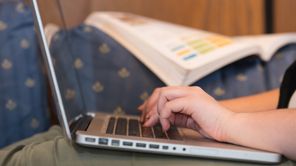 Close-up of a person typing on a laptop with an open textbook in the background.