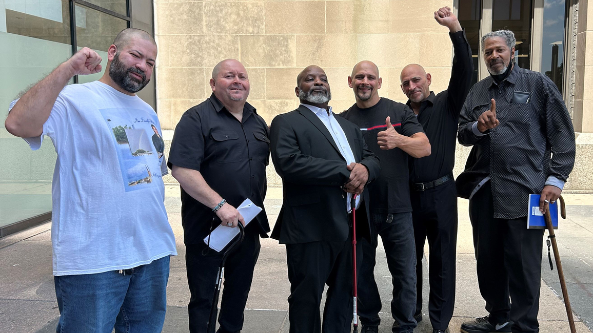 Six men stand smiling outside a courthouse, some raising fists. Left to right: Daniel Rodriguez, Fernando Gomez, Michael McCastle, Gregorio Cardona, Robert Cardona, Lowell Higgins-Bey.