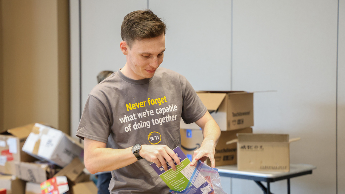 A person packing a colorful bag with supplies in a room filled with boxes. Their shirt reads, "Never forget what we're capable of doing together."