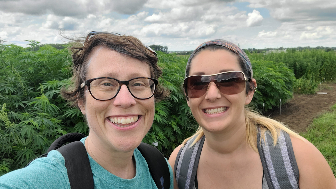 Two smiling people outdoors, cloudy sky, green foliage in the background.