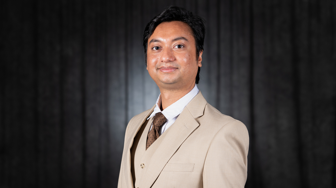 A man in a beige suit and tie, smiling, against a dark background.
