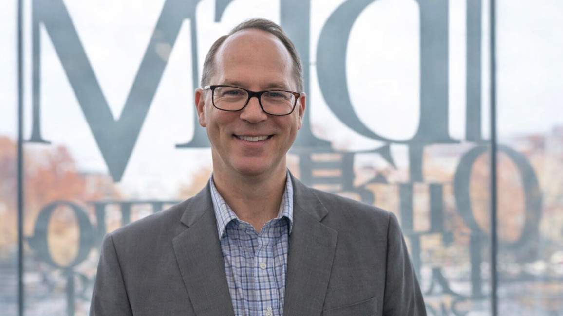 Smiling man in suit standing in front of blurred window.