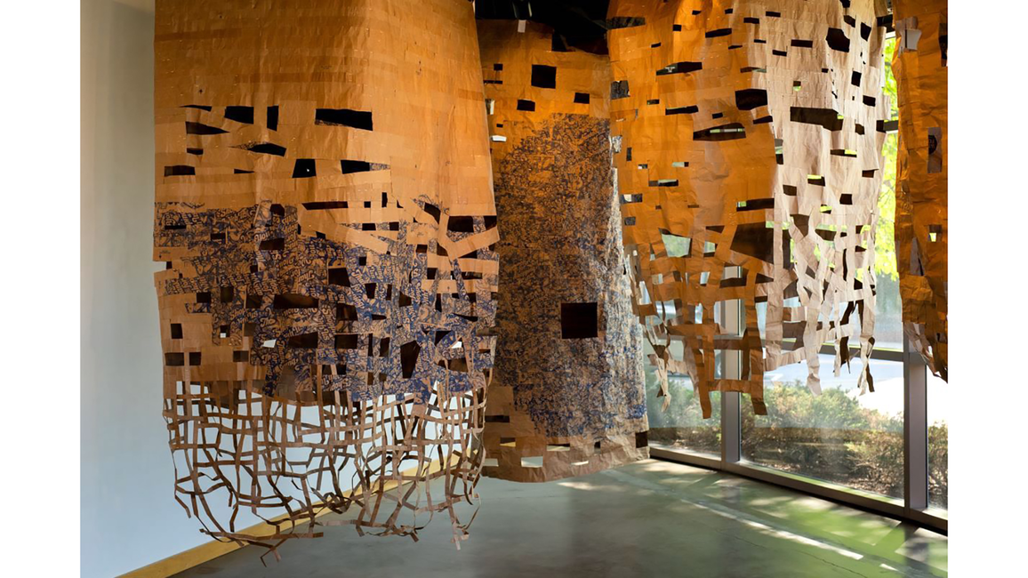 Hanging woven art installation with cutouts, warm tones, near windows.