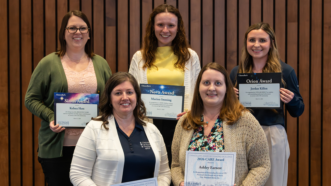 Five women smiling, each holding a certificate, standing against a wooden wall.