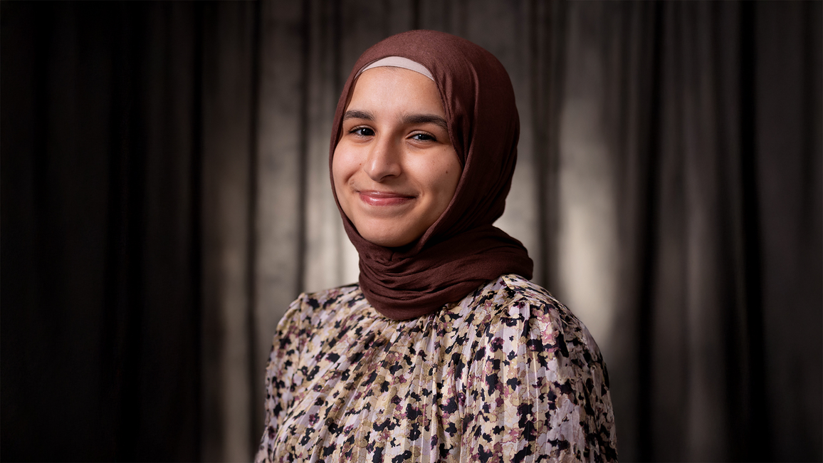 Smiling person in a floral blouse and dark hijab in front of a grey background.