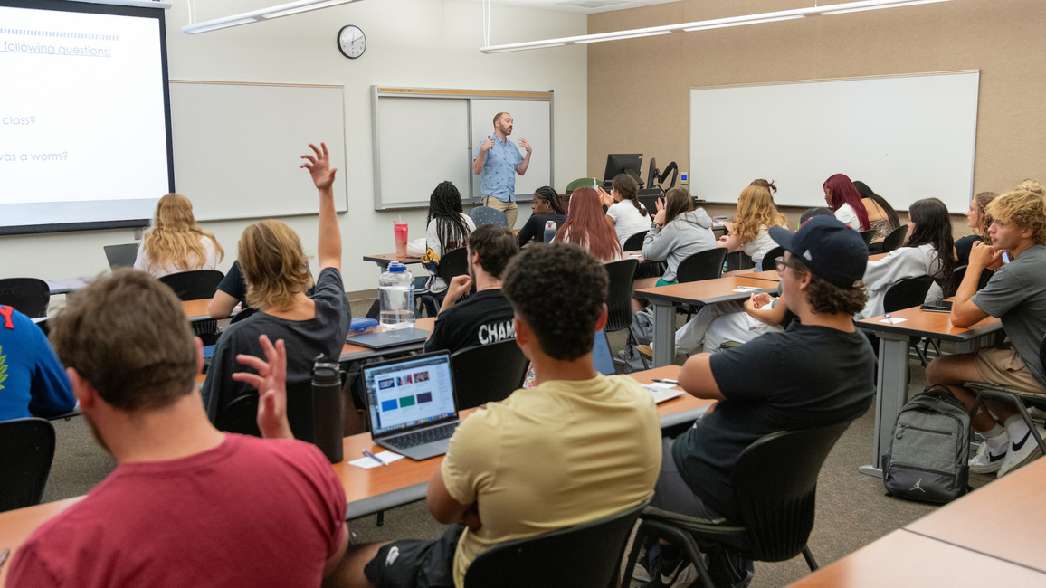 Classroom with students seated, one raising a hand. Instructor at the front talking.