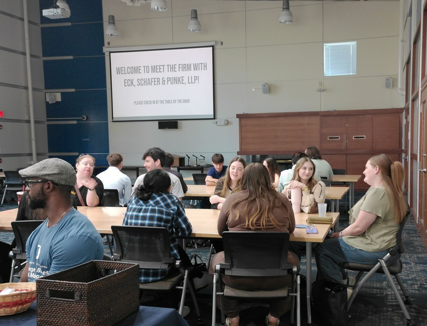 Meeting room with people seated at tables, screen displaying text.