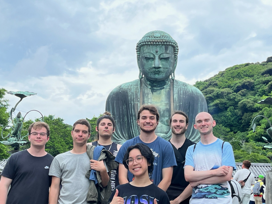 Seven people smiling in front of a large Buddha statue under a cloudy sky.