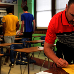 a high school teacher filling out a form at their desk while his students look on