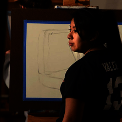an art student looking at a still life subject while drawing it on her canvas on an easel