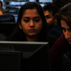 a professor standing next to a student at a computer desk showing the student something on a computer
