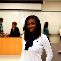 a female student smiling in a classroom setting with a professor and students in the background