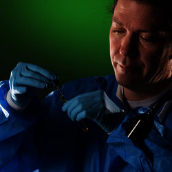 Scientist in blue protective gear under green lighting using a dropped to add liquid in beaker.