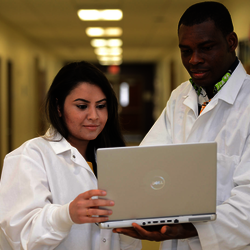 two healthcare informatics students in lab coats looking at a laptop in a hallway