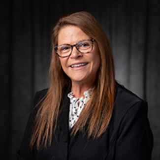 Smiling woman in glasses with long hair, wearing a dark blazer against a dark background.