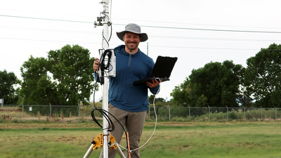 Man with hat holding a laptop, standing by equipment in a grassy outdoor setting.