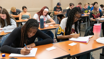 Students attentively writing in a classroom, sitting at desks with notebooks.