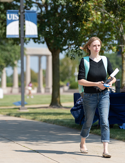 A student carrying books walking on a sidewalk with the colonnade in the background.