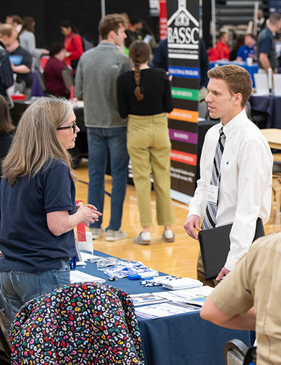 A job fair attendee wearing business attire speaks with a representative at an informational table.