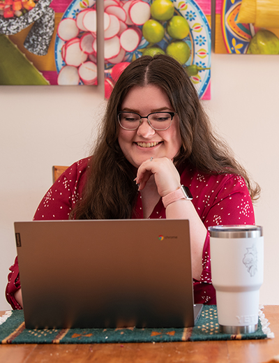 A person in glasses, wearing a red shirt, smiles while working on a laptop at a table with a white travel mug nearby.