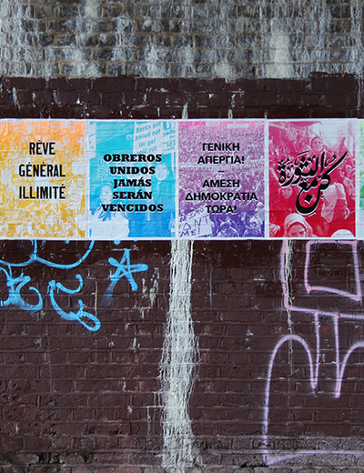 A vibrant poster series on a graffiti-covered brick wall. Each colorful panel displays multilingual text.