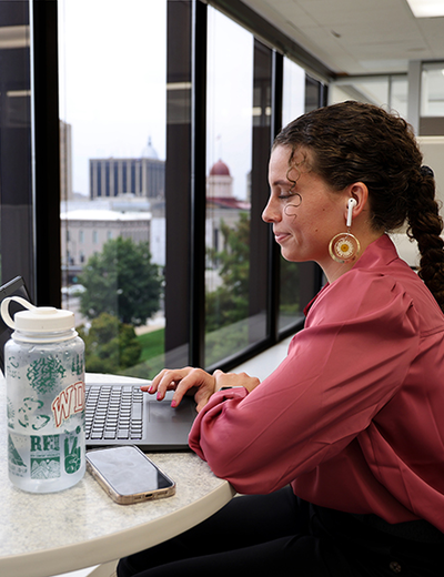Young woman works on a laptop at a high-rise office window with earbuds, water bottle, and phone nearby.
