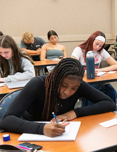 Students sit at desks in a classroom, writing notes and focusing on their work.
