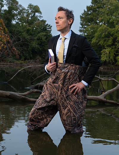 Brandon Derman stands in water wearing waders and a suit, holding a book, with trees in the background.