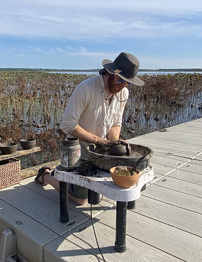 A person shapes clay on a pottery wheel outdoors on a dock at the Emiquon Preserve, surrounded by wetland plants and water.