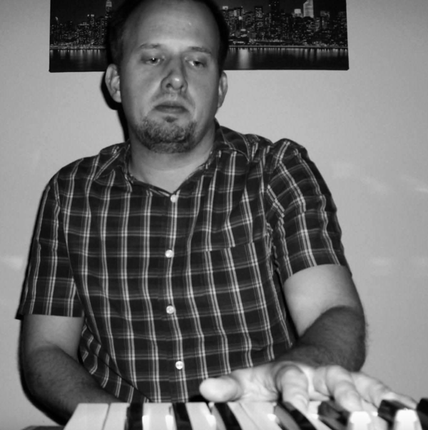 Man in plaid shirt playing keyboard in dim light, looking focused.