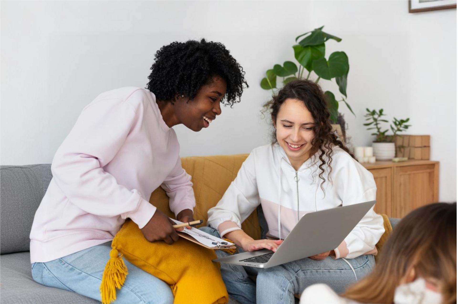 Two young women collaborating on a project, one holding a clipboard and the other using a laptop, sitting on a couch in a cozy living room