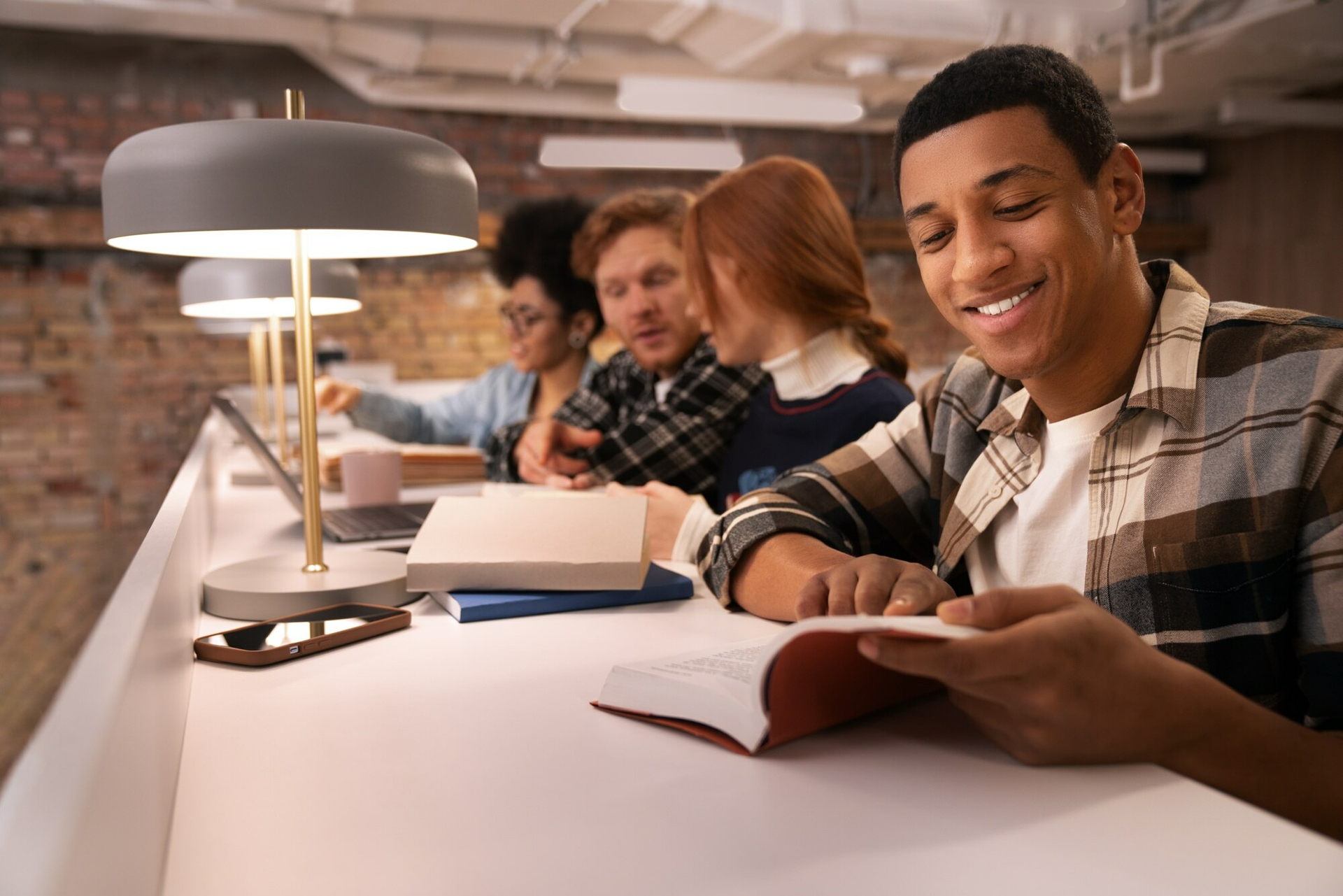 A group of students studying together at a communal table in a modern library. The student in the foreground is smiling and reading, while others are engaged in discussions and using laptops