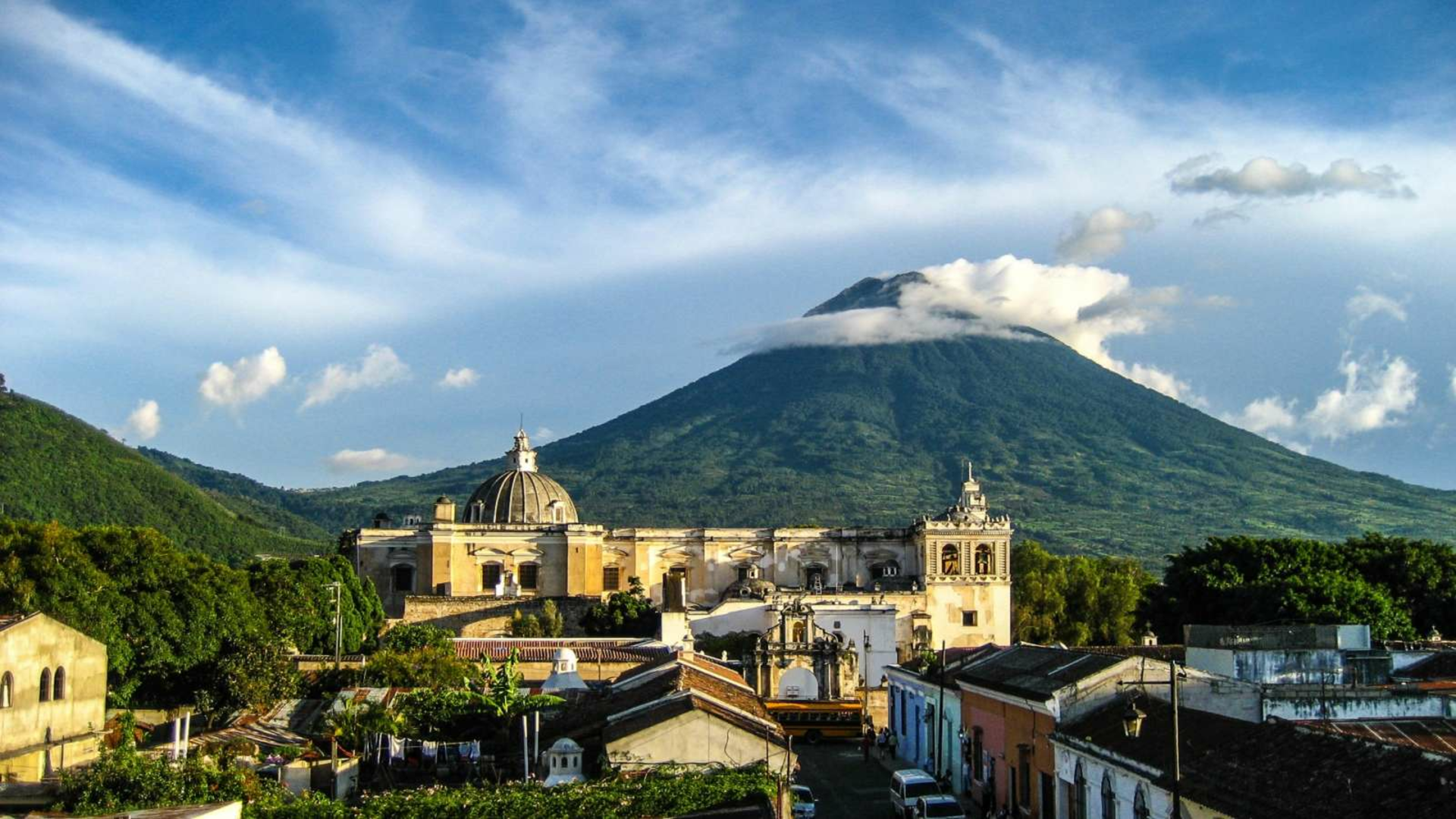 Image is a picture taken from a high view in Antigua, Guatemala. There is a blue sky with one large volcano toward the back of the image. It is covered in foliage, making it green. In front and below of the volcano show buildings and structures. There is a line of brightly covered buildings toward the front of the image. Farther back and higher up is a large building that is a cream color.