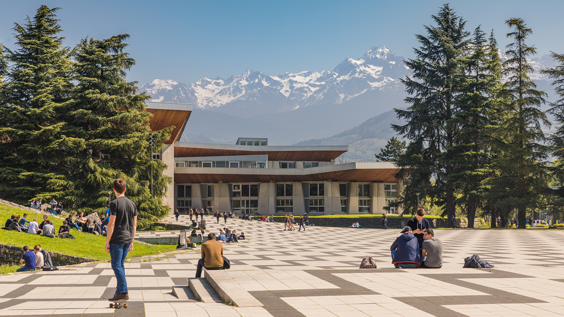 Part of the campus that has chevron-patterned pavement. There are several students sitting and walking on the pavement. To the left is a grassy lawn with students sitting on it. Further back, is a campus building with large pine trees on either side of it. In the background is a picture of the French Alps, a mountain range with snow on it.