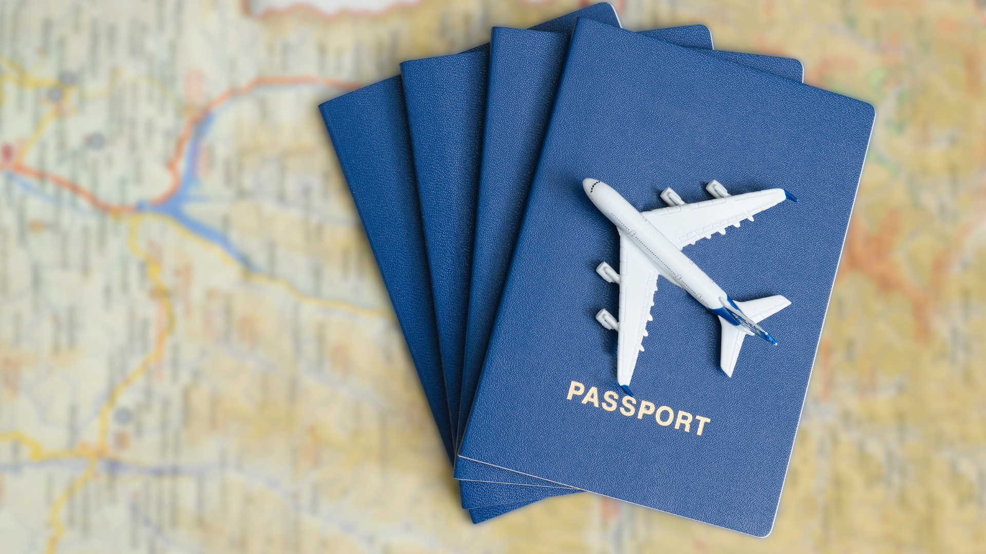 A colorful map that is out of focus and the backdrop. Laying on the map are several passports stacked on-top of each other in fanned-out pattern. Laying on top of the passports is a white toy plane.