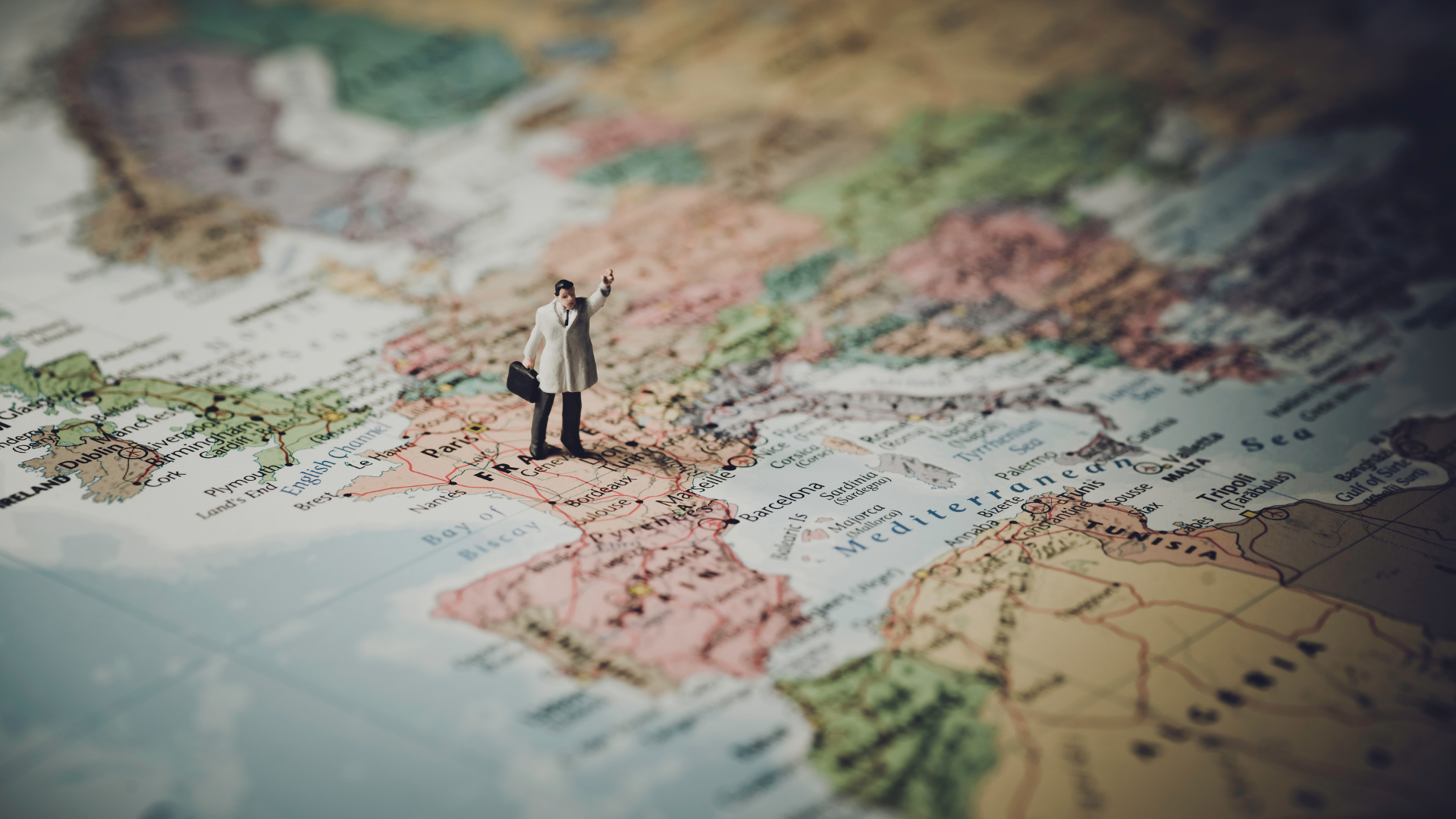 Part of a world map with a small plastic figurine standing on it. The figurine is holding a briefcase, wearing a hat, trench coat, and black pants and shoes.