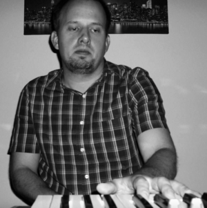 Man in plaid shirt playing keyboard in dim light, looking focused.